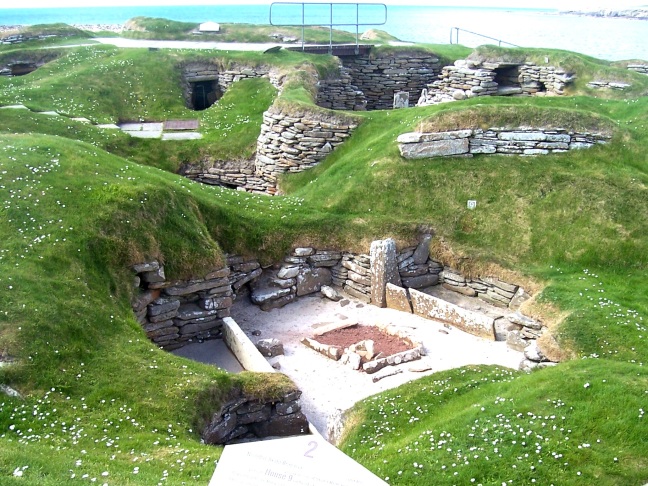 A home at Skara Brae without out its earthen roof and walls. Hearth in the middle. Sleep area beyond. Other homes in the background.